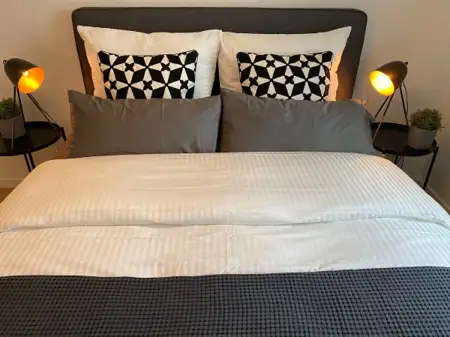 Gemütliches Bett mit gestreiften Bettlaken, dekorativen Kissen und modernen Nachttischlampen.